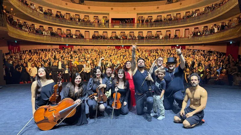 Músicos com violinos e violoncelo reunidos no palco, com a plateia do teatro ao fundo