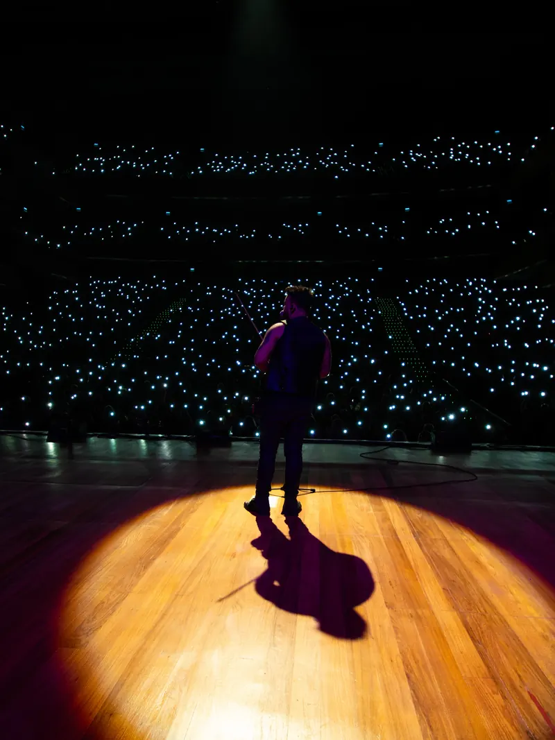 Músico sozinho sob um holofote no palco de madeira, de frente para uma plateia iluminada por luzes de celulares